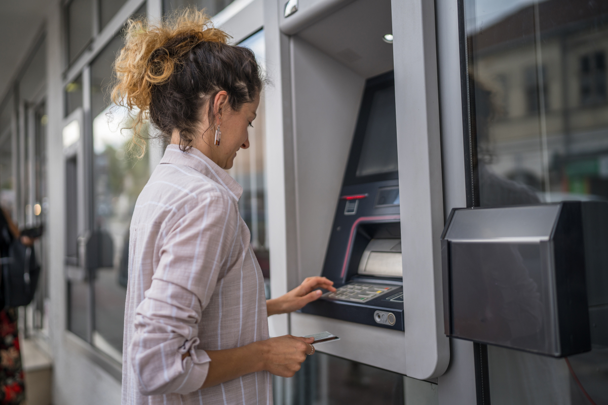 Kobieta stoi przed bankomatem, tuż przed włożeniem do niego karty płatniczej