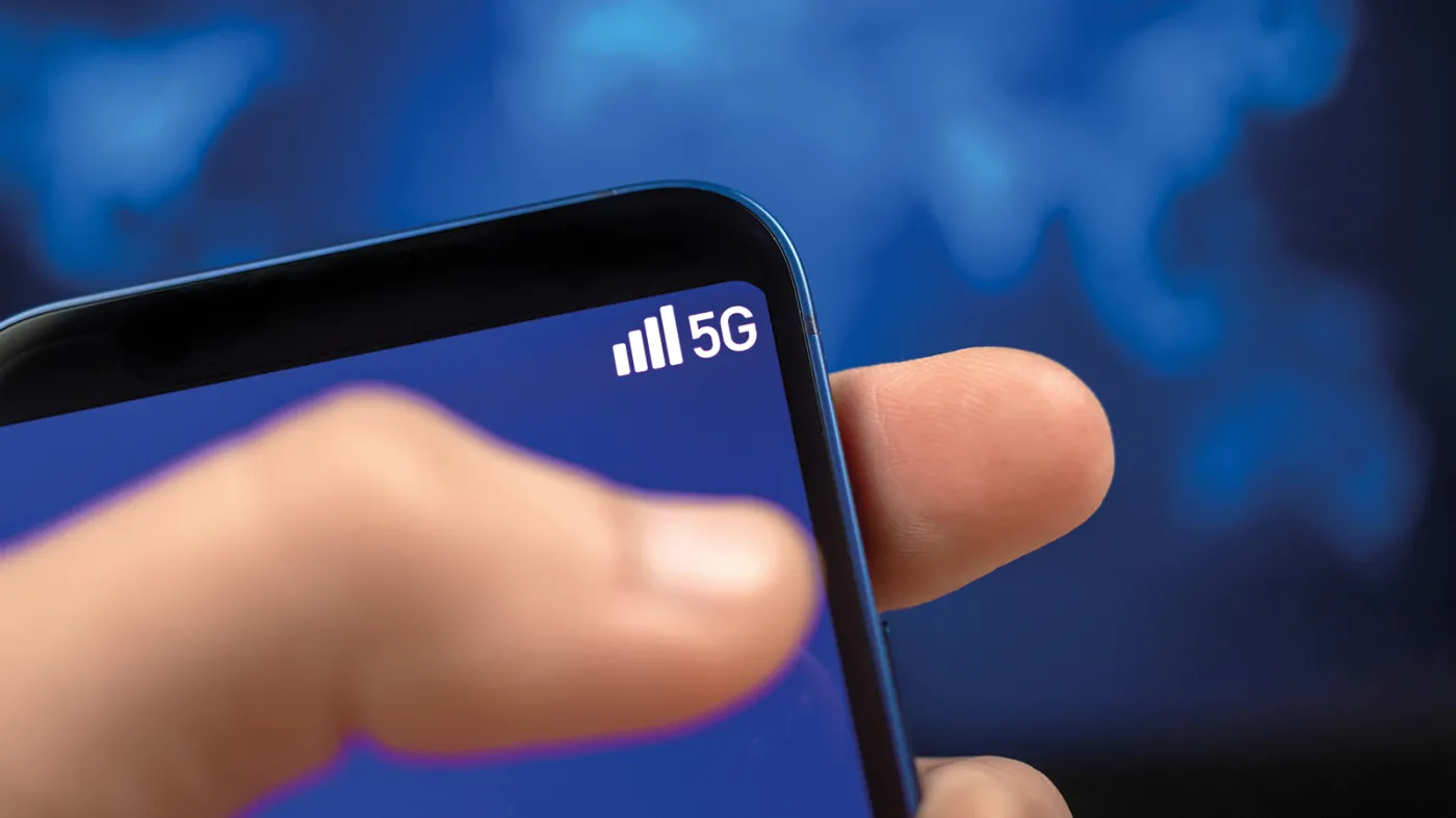 Na pierwszym planie dłoń ze smartfonem pokazującym niewielki symbol 5G