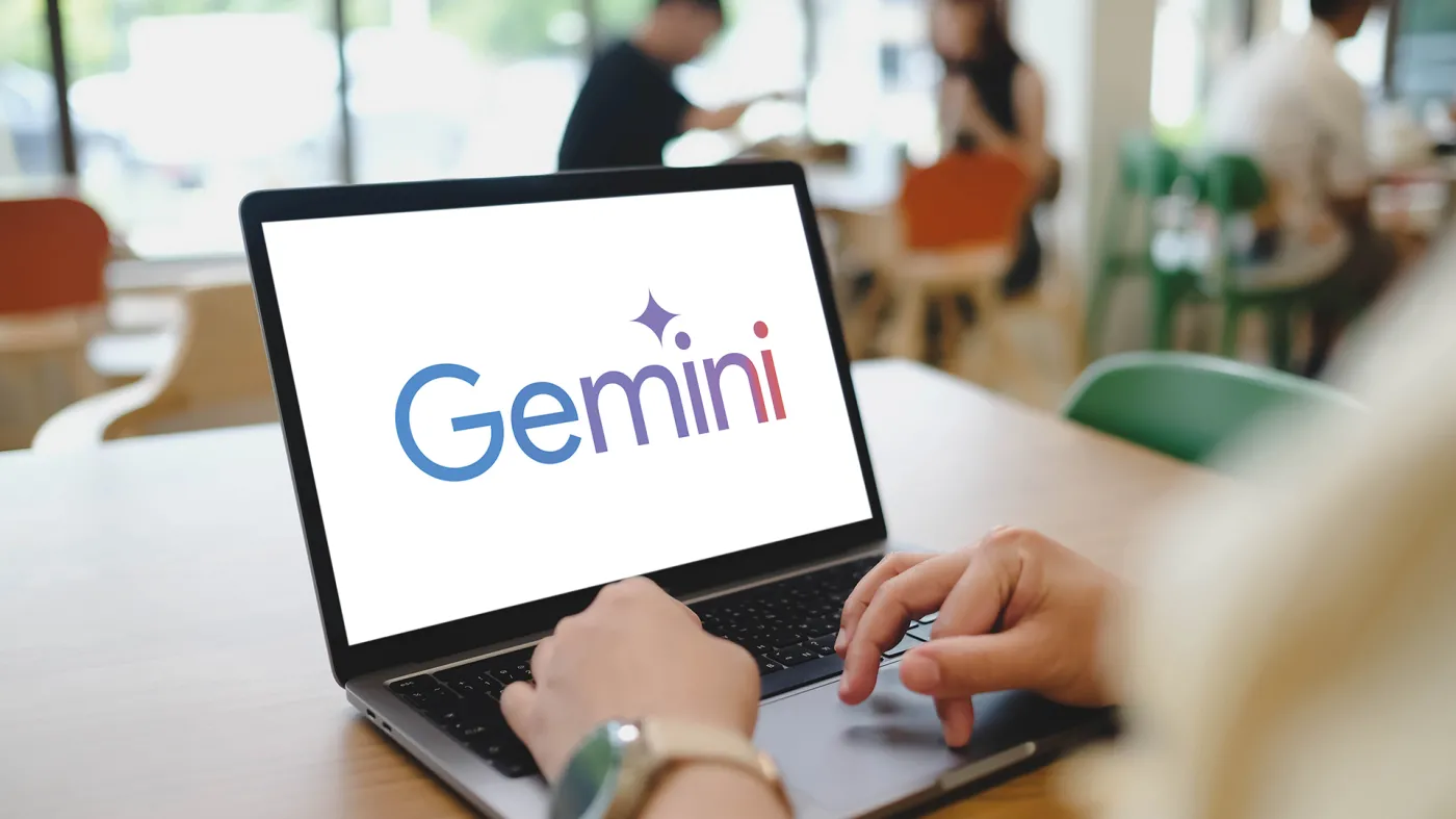 Młoda osoba (student/studentka) w środowisku uczelnianym przy biurku z laptopem. Na ekranie widoczne jest czytelne logo Google Gemini