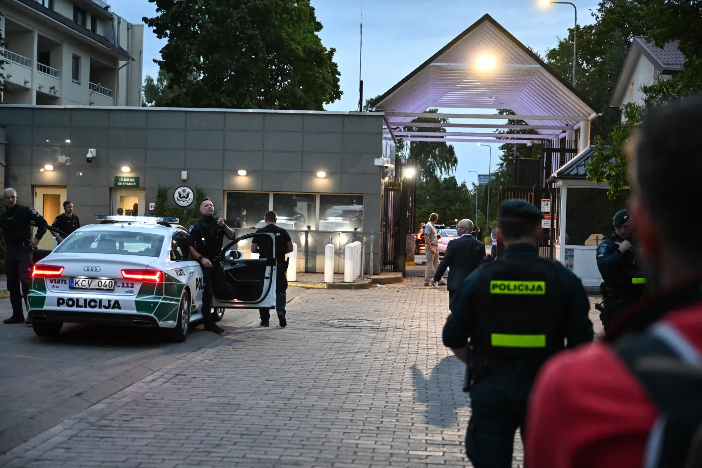 Przyjazd uwolnionych więźniów z Białorusi. Na zdjęciu widać grupę policjantów stojących przed budynkiem z szarą elewacją i zadaszoną bramą wjazdową. Na pierwszym planie znajduje się policyjny radiowóz z napisem „Policija” i otwartymi drzwiami.
