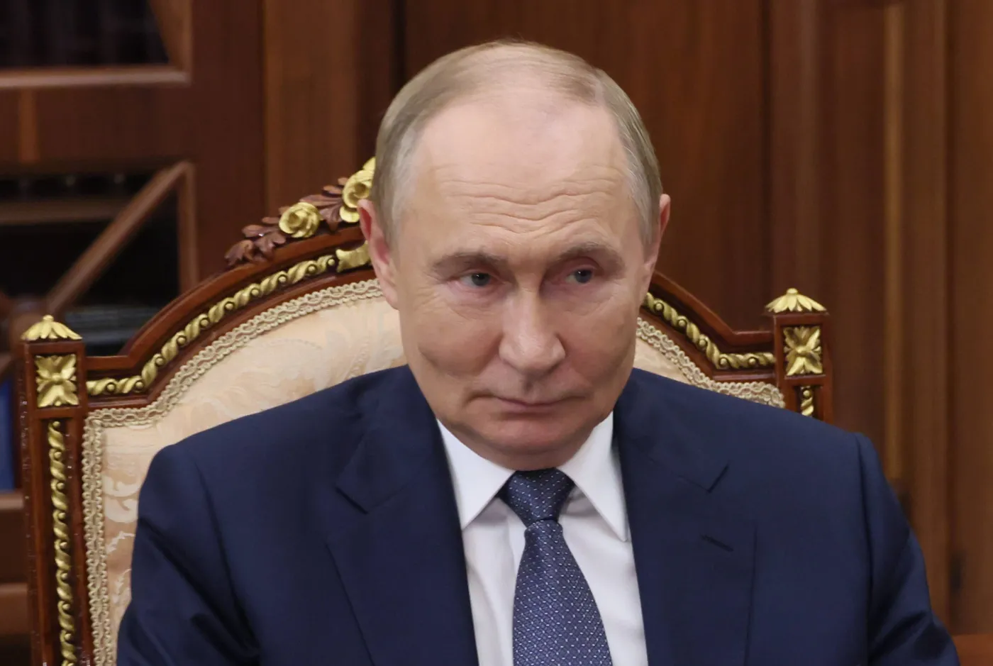 Władimir Putin w garniturze siedzi na ozdobnym, jasnym fotelu z drewnianymi zdobieniami, patrząc poważnym wzrokiem w bok