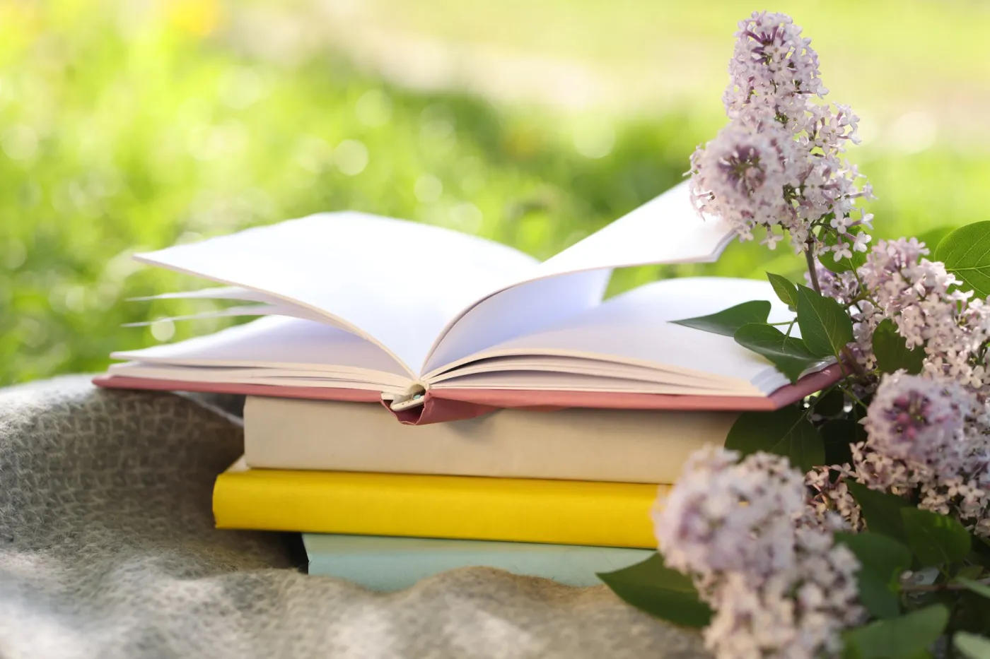 Stacked,Books,,Lilac,Flowers,And,Blanket,On,Green,Grass,Outdoors,