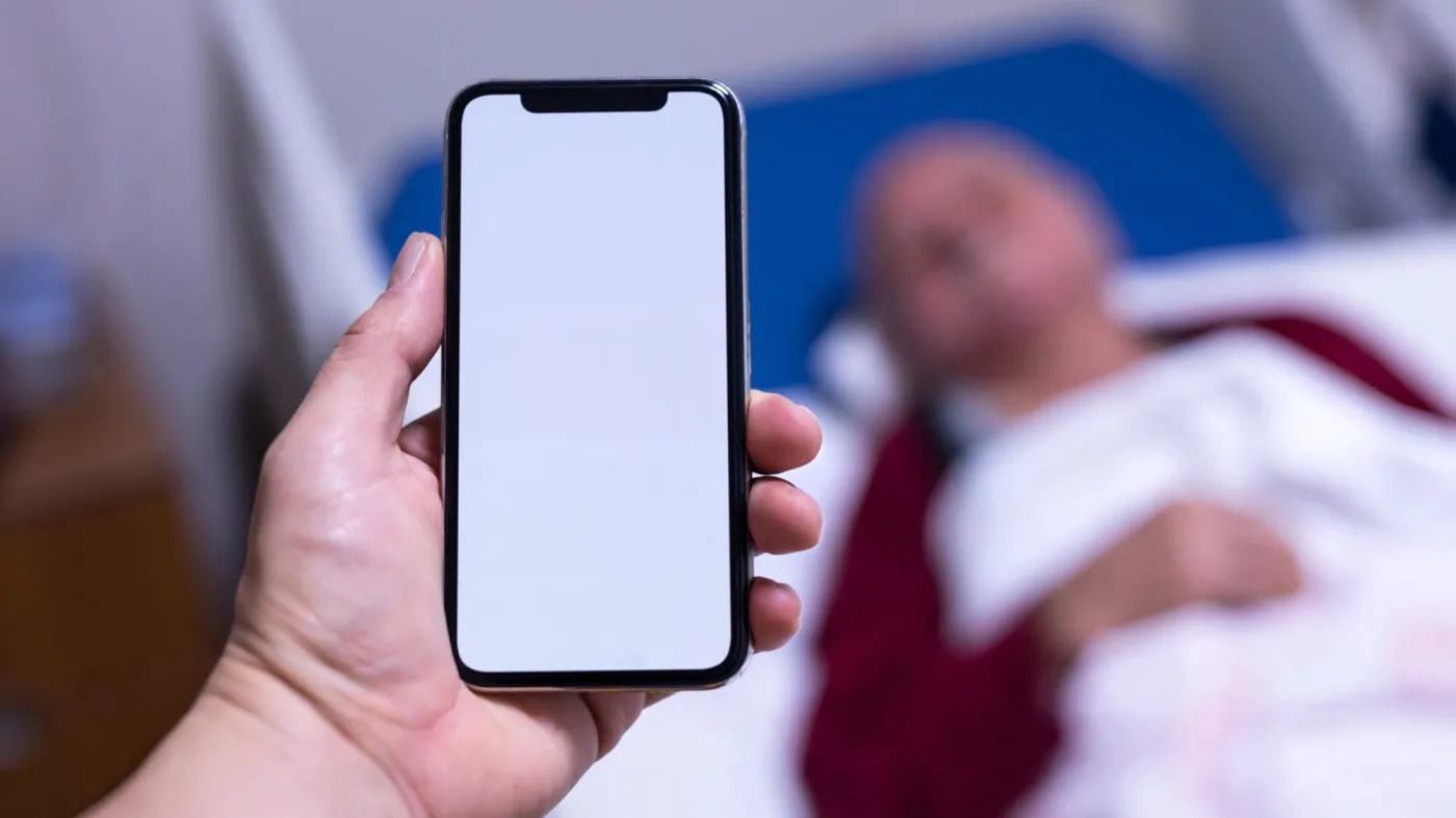 Na pierwszym planie osoba w wyciągniętej dłoni trzyma smartfon z włączonym, białym ekranem, na drugim planie w łóżku szpitalnym leży pacjent.