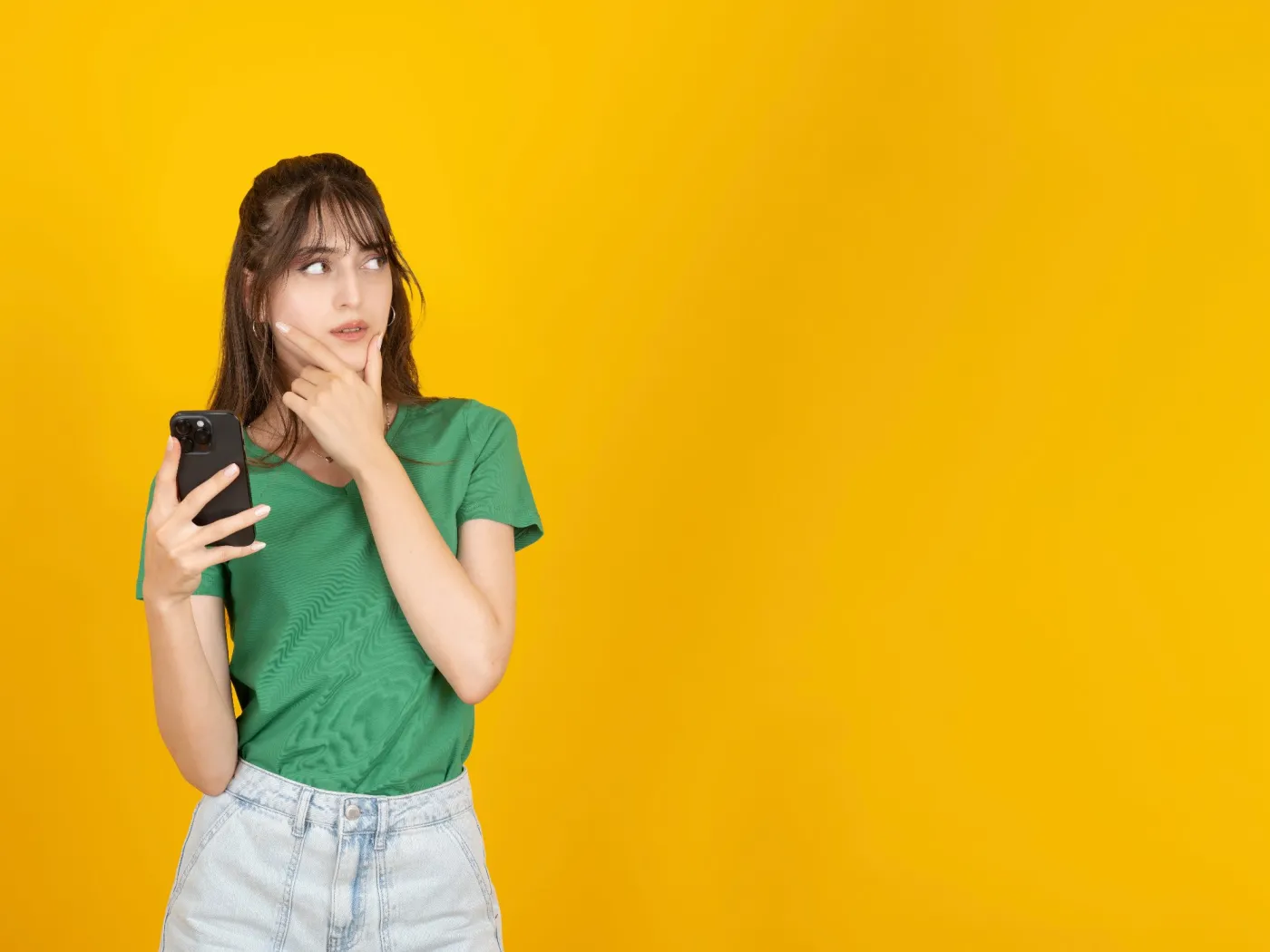 Young,Woman,Holding,Smartphone,While,Touching,Chin,Thoughtfully,,Wearing,Green