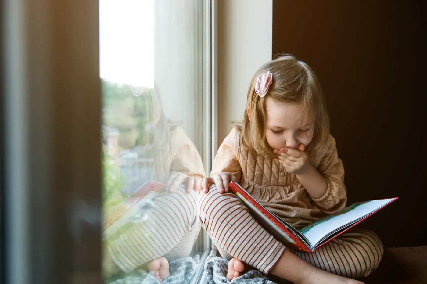 Little,Girl,Is,Reading,A,Book,While,Sitting,Near,Window
