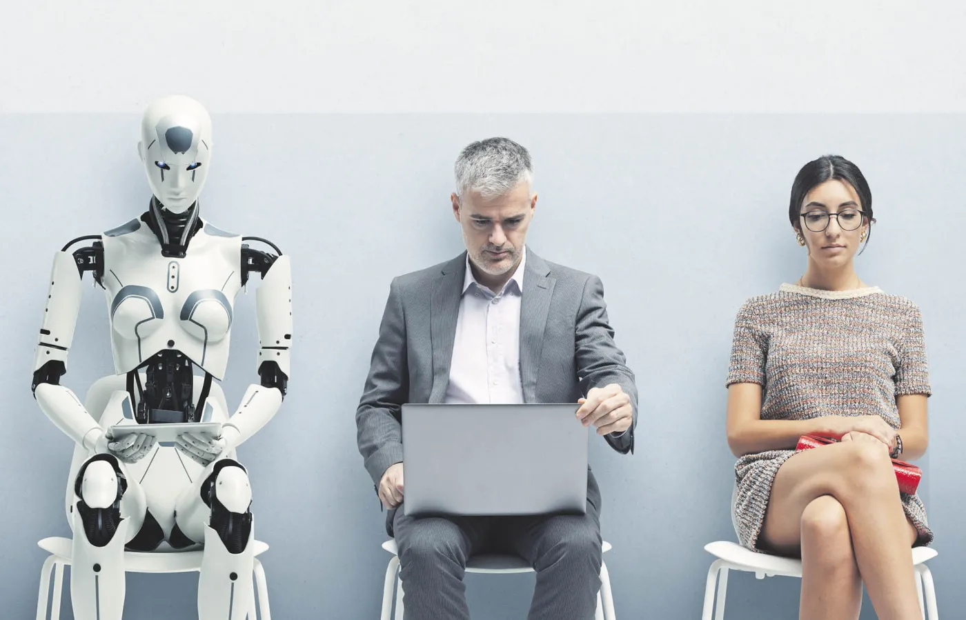 Pomieszczenie sugerujące poczekalnię przed rozmową o pracę. Na dwóch krzesłach siedzą ludzie, a na jednym z nich humanoidalny robot.