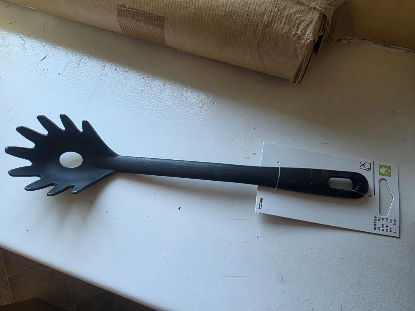 Czarna, plastikowa łyżka do spaghetti z ząbkami i centralnym otworem, leżąca na białym blacie. Do uchwytu wciąż przymocowana jest papierowa metka z kodem kreskowym i logotypami, w tym ikoną kieliszka i widelca. W tle widać fragment kartonowego opakowania