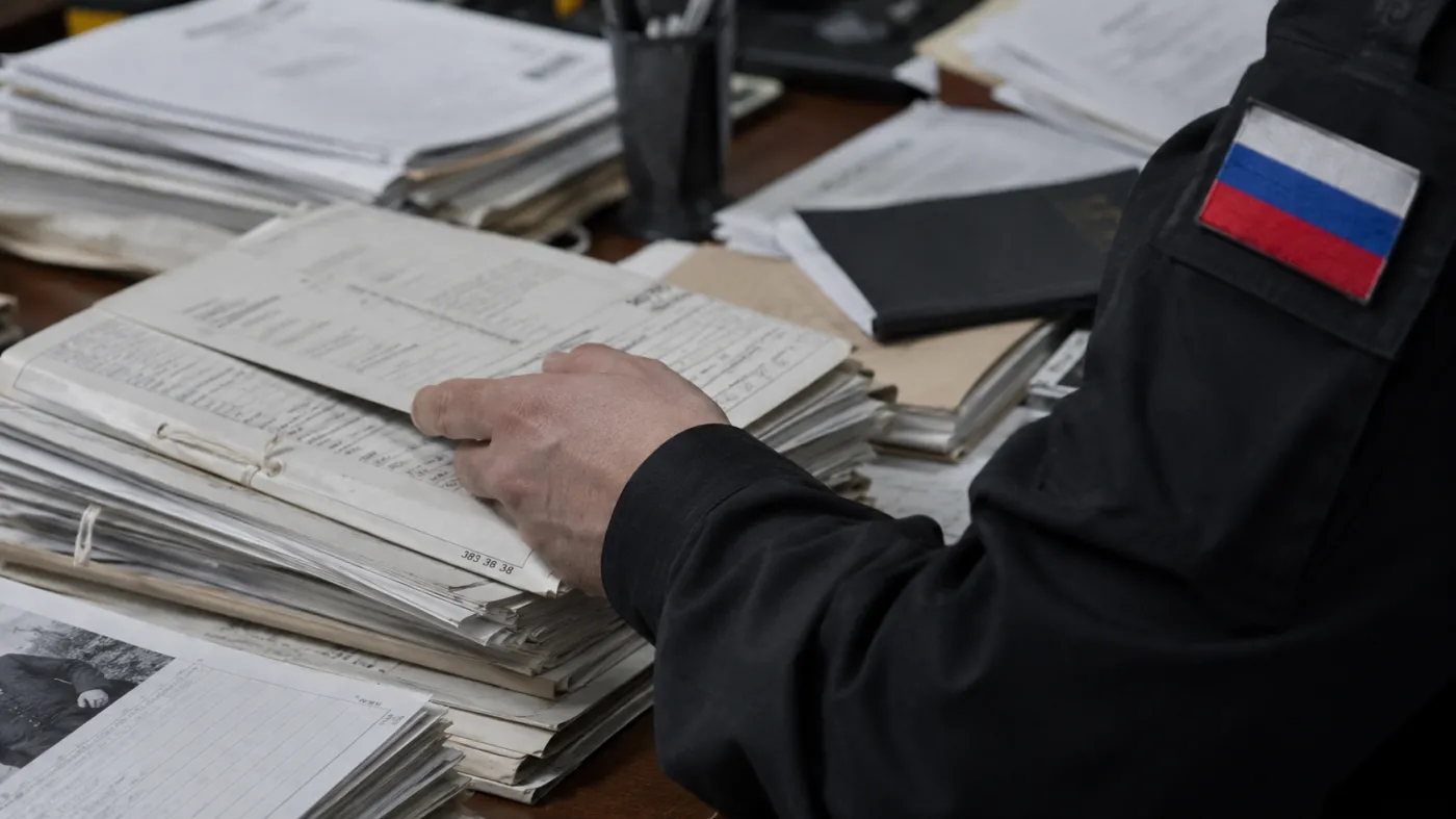 zdjęcie poglądowe przedstawiające biurko zasypane papierowymi aktami i dokumentami. Na pierwszym planie widać rękę mężczyzny w ciemnym mundurze, przeglądającego teczki; na jego ramieniu znajduje się naszywka w barwach rosyjskiej flagi. Scena sugeruje pracę służb lub analizę dokumentów.