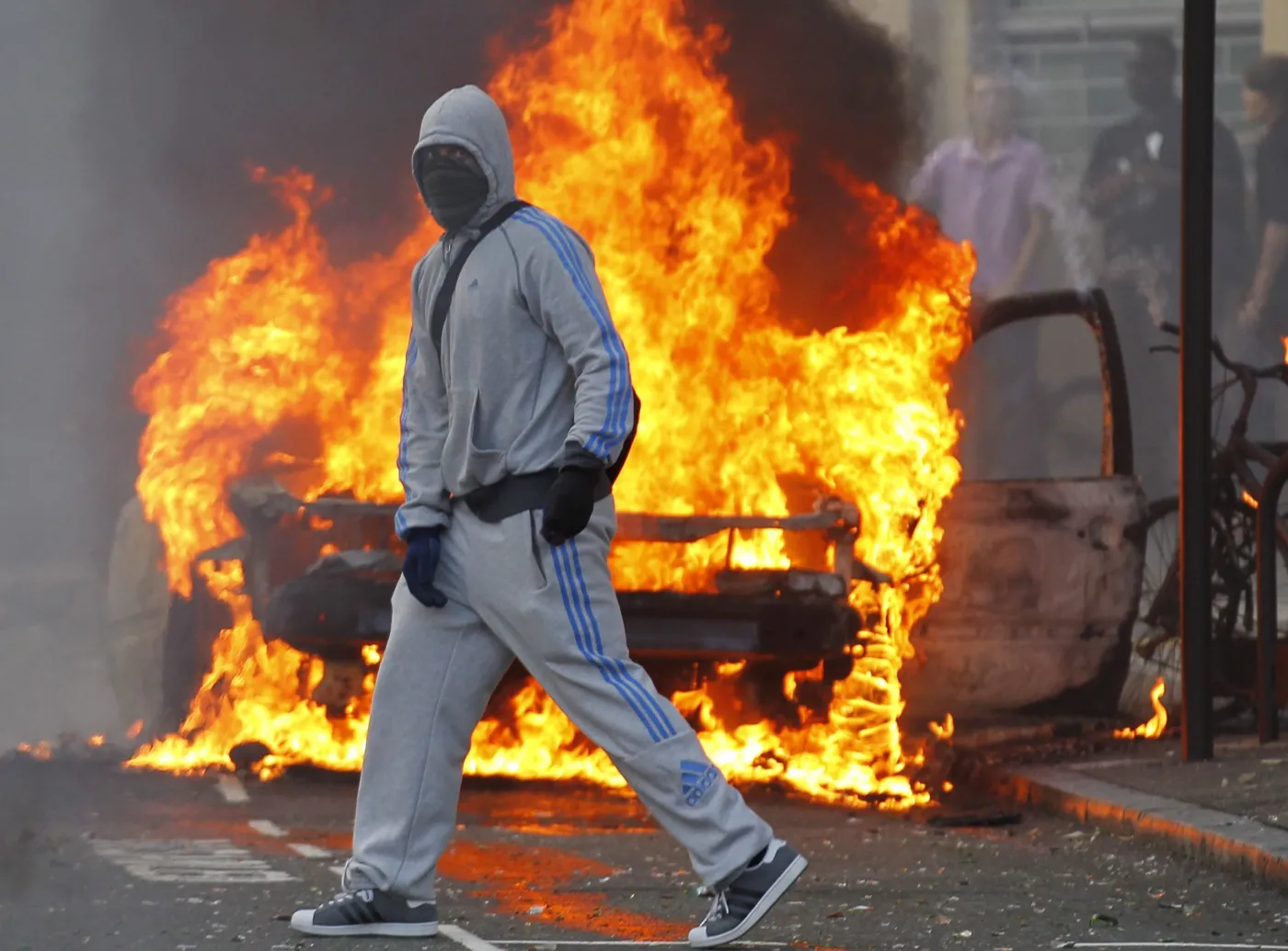 Jeden z zamaskowanych chuliganów przed płonącym samochodem w Hackney