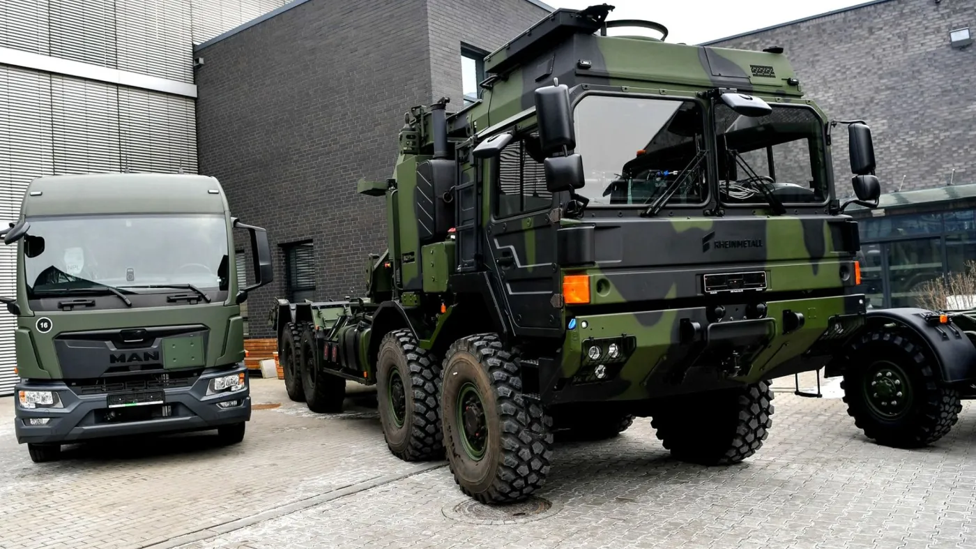 Dwie zielone ciężarówki wojskowe w kamuflażu – po prawej ciężarówka Rheinmetall, po lewej pojazd MAN, zaparkowane przed nowoczesnym budynkiem