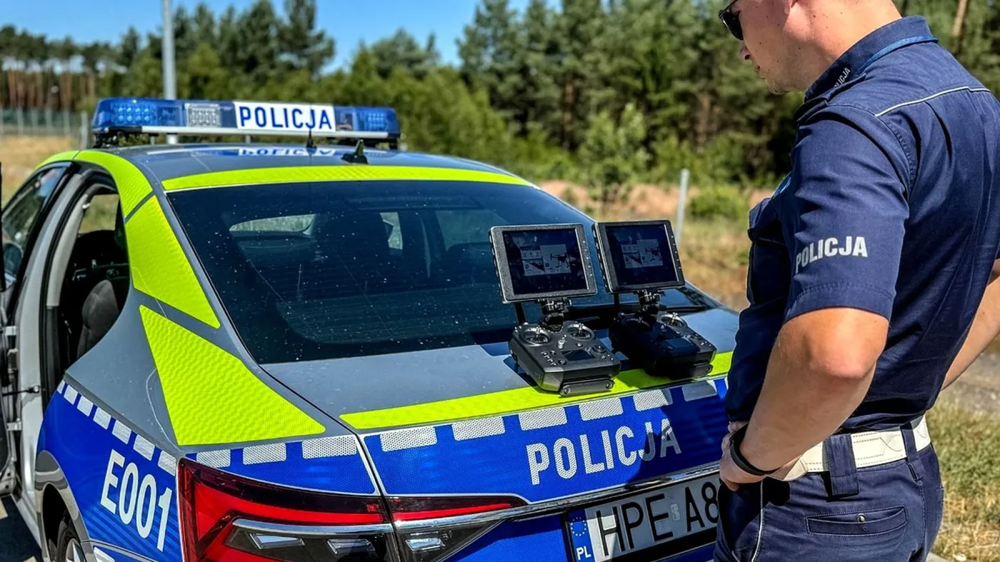 Umundurowany policjant stojący przy nowym, srebrno-niebiesko-żółtym radiowozie i patrzący na dwa ekrany kontrolerów do drona leżące na klapie bagażnika samochodu