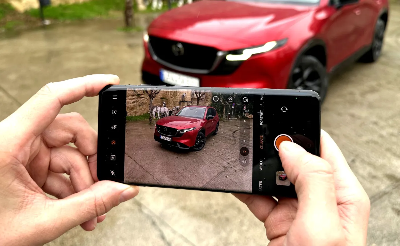 Osoba trzymająca smartfon i robiąca zdjęcie czerwonego samochodu na parkingu, na ekranie telefonu widoczne menu aparatu w języku polskim, ilustracja do artykułu o kontrolach abonamentu RTV w autach