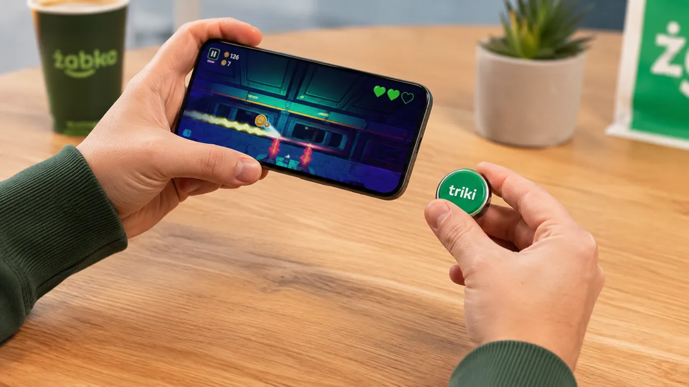 Użytkownik trzyma w dłoni smartfon z uruchomioną grą mobilną oraz miniaturowy kontroler Triki przypominający kapsel. Urządzenie połączone jest z telefonem i służy do sterowania grą za pomocą ruchów dłoni. Na stole widoczne są elementy związane z marką Żabka.