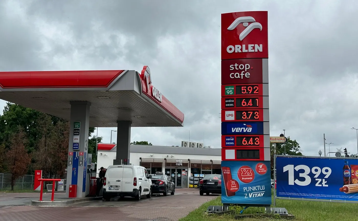 Czerwony pylon stacji Orlen i stanowiska kasowe stop cafe. Ceny na pylonie: benzyna 95 za 5,92 zł, ON za 6,41 zł, LPG za 3,73 zł. Obok stoi biały samochód dostawczy