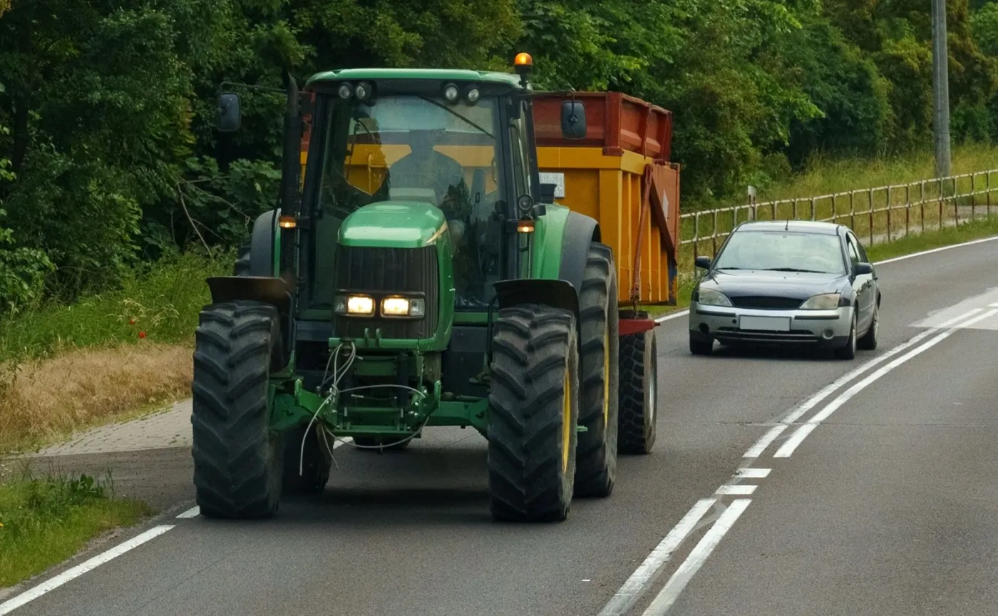Zielony traktor z pomarańczową przyczepą jadący drogą w Polsce, za którym jedzie srebrny samochód osobowy tuż przy linii podwójnej ciągłej