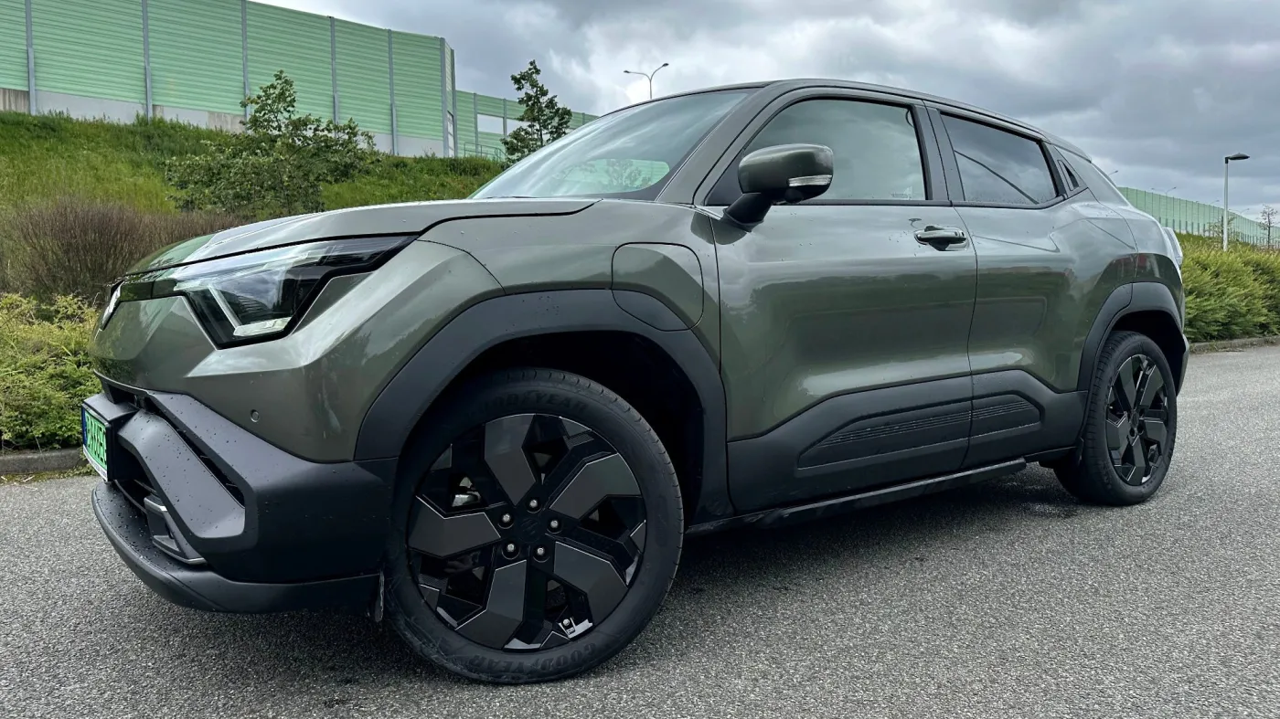 Nowy, oliwkowozielony elektryczny SUV Suzuki e-Vitara z czarnym dachem, ujęcie z przodu pod kątem, zaparkowany na drodze pod zachmurzonym niebem