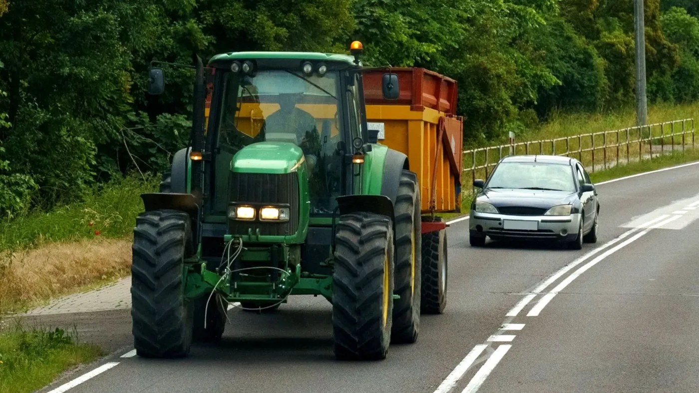 Zielony traktor z pomarańczową przyczepą jadący drogą w Polsce, za którym jedzie srebrny samochód osobowy tuż przy podwójnej linii ciągłej