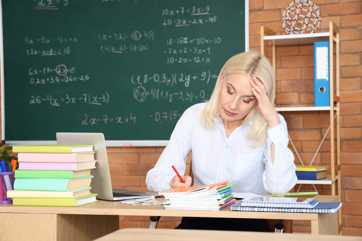 Tired,Female,Math,Teacher,Checking,Copybook,At,Table,In,Classroom
