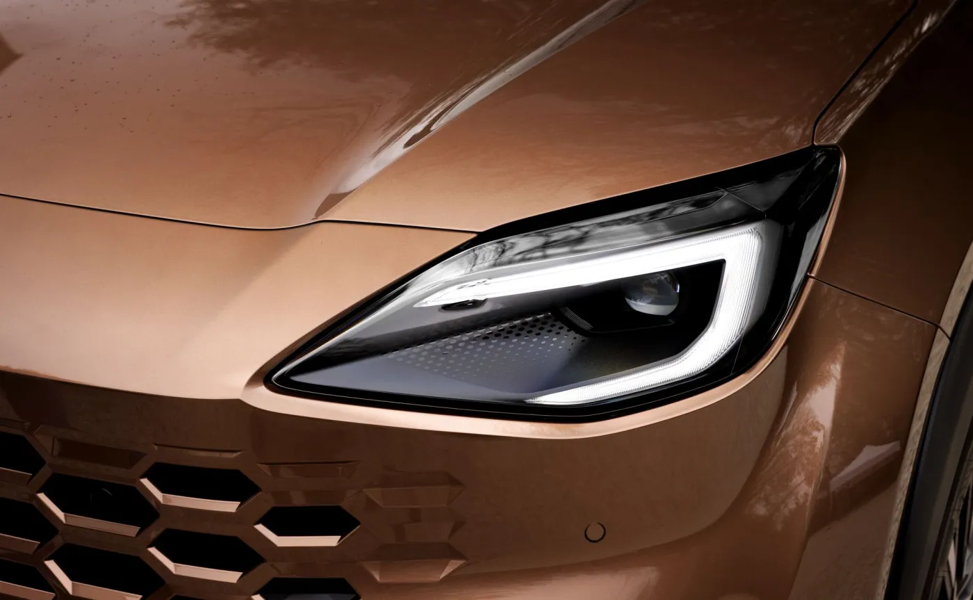 Zbliżenie na nowoczesny reflektor LED i grill o strukturze plastra miodu nowej Toyoty Yaris Cross w miedziano-brązowym lakierze Precious Bronze, z kropelkami deszczu na masce