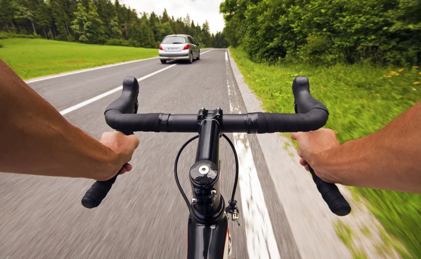 Perspektywa z kierownicy roweru szosowego (dłonie kolarza na baranku) jadącego po asfalcie. Przed nim, w odległości kilku metrów, jedzie srebrny samochód osobowy. Po prawej stronie widoczna jest ciągła linia krawędziowa i zielone pobocze z drzewami. Sytuacja drogowa ilustruje dylemat zachowania bezpiecznego 1 metra odstępu przy wyprzedzaniu rowerzysty