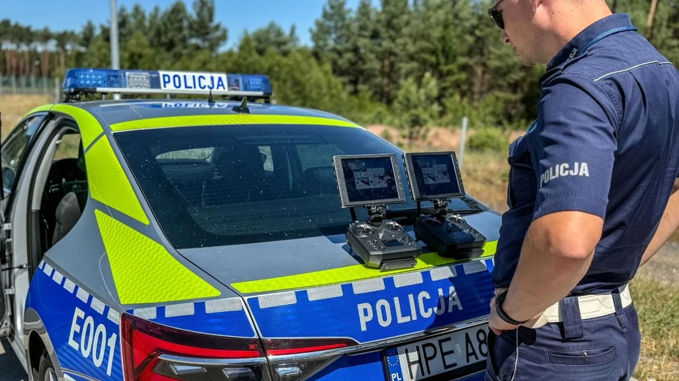 Umundurowany policjant stojący przy nowym, srebrno-niebiesko-żółtym radiowozie i patrzący na dwa ekrany kontrolerów do drona leżące na klapie bagażnika samochodu