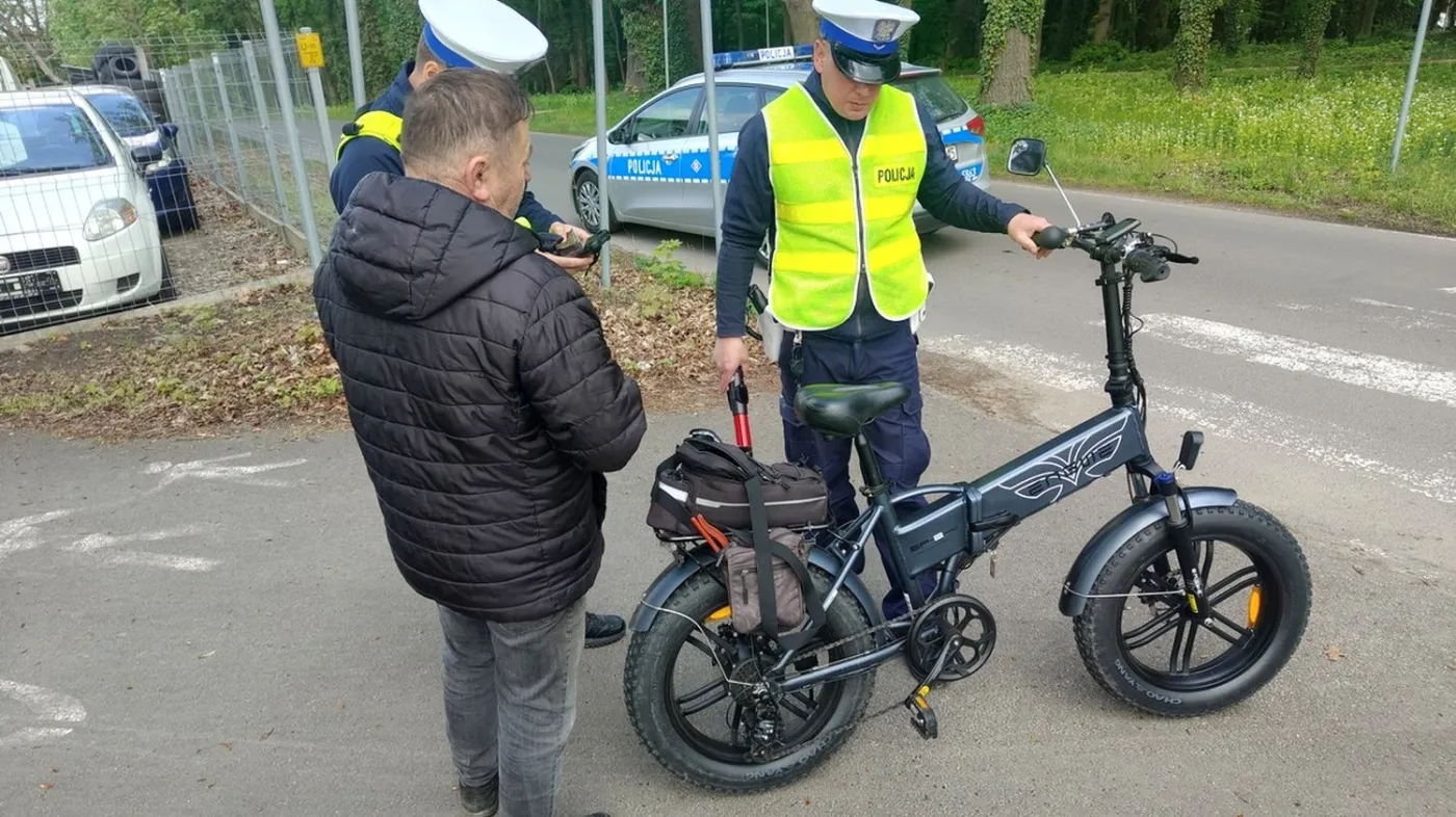 Kontrola drogowa policji. Dwóch policjantów drogówki w żółtych kamizelkach odblaskowych i białych czapkach sprawdza mężczyznę z czarnym, składanym rowerem elektrycznym o grubych oponach. W tle widoczny radiowóz