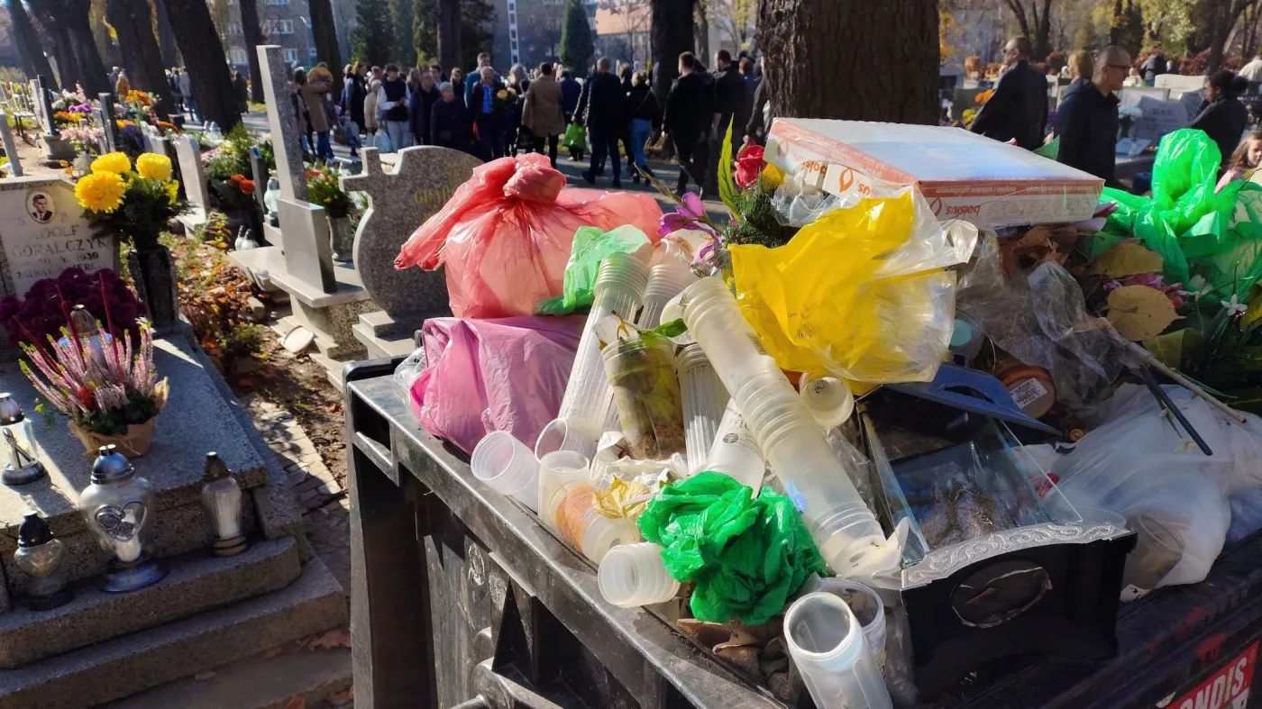 Śmieci wystają z kontenera na odpady na cmentarzu - w tle groby i przechodzący ludzie