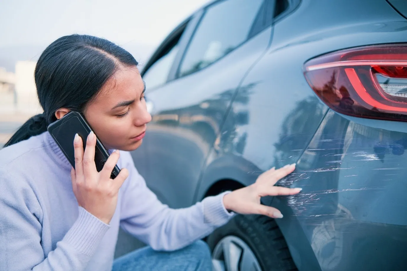 Kobieta oglądająca zarysowanie na zderzaku samochodu i rozmawiająca przez telefon