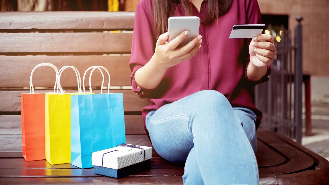 Kobieta trzymająca kartę płatniczą i telefon, obok kolorowe torby i paczka