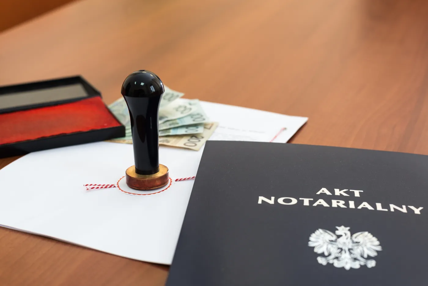 Rozłożone na stole dokumenty, teczka opisana jako "Akt notarialny", stempel oraz pieniądze
