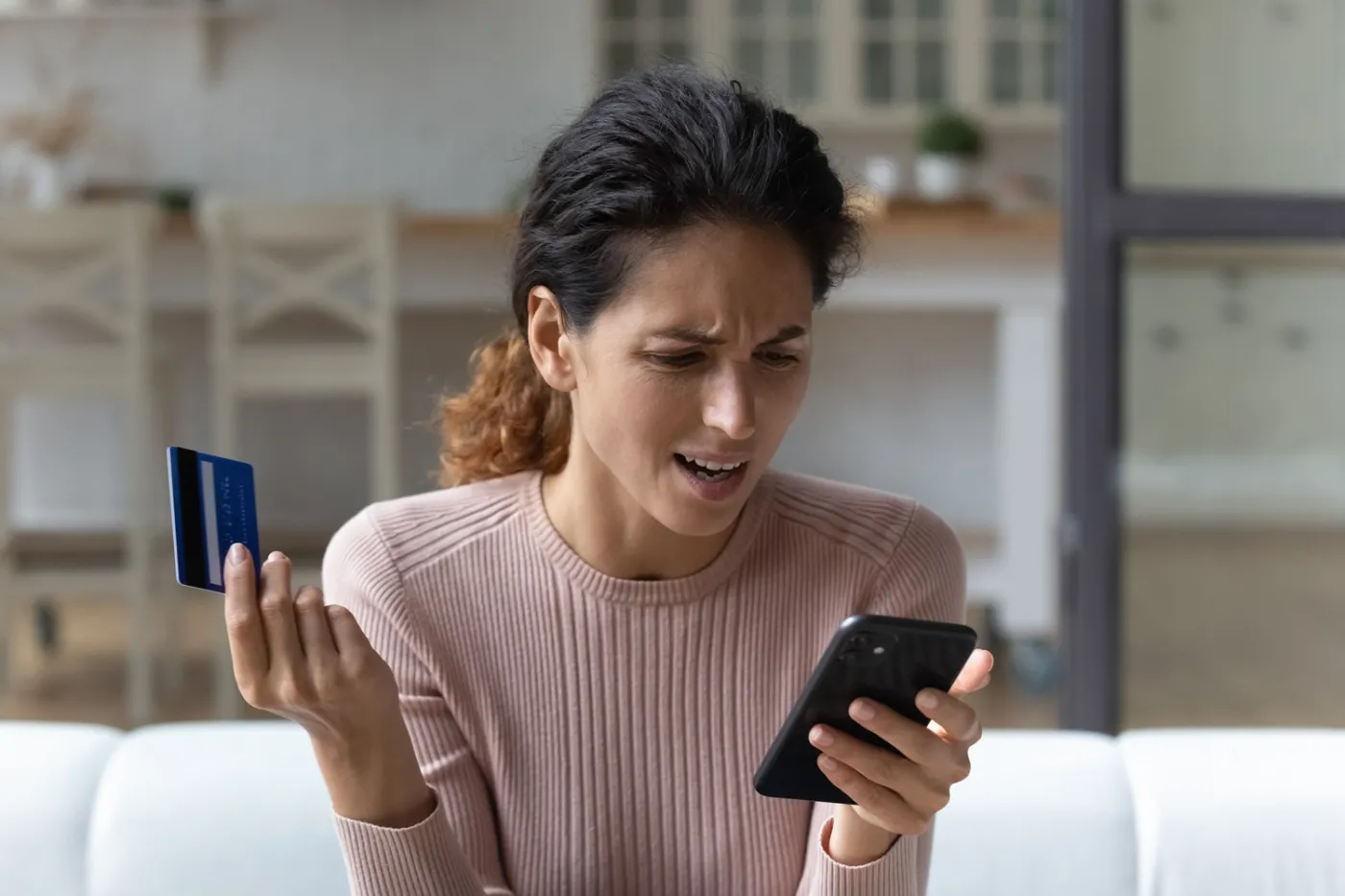 Kobieta siedzi na sofie w salonie: w jednej dłoni trzyma smartfon, na który patrzy, a w drugiej kartę. Jest zszokowana tym, co widzi w telefonie.