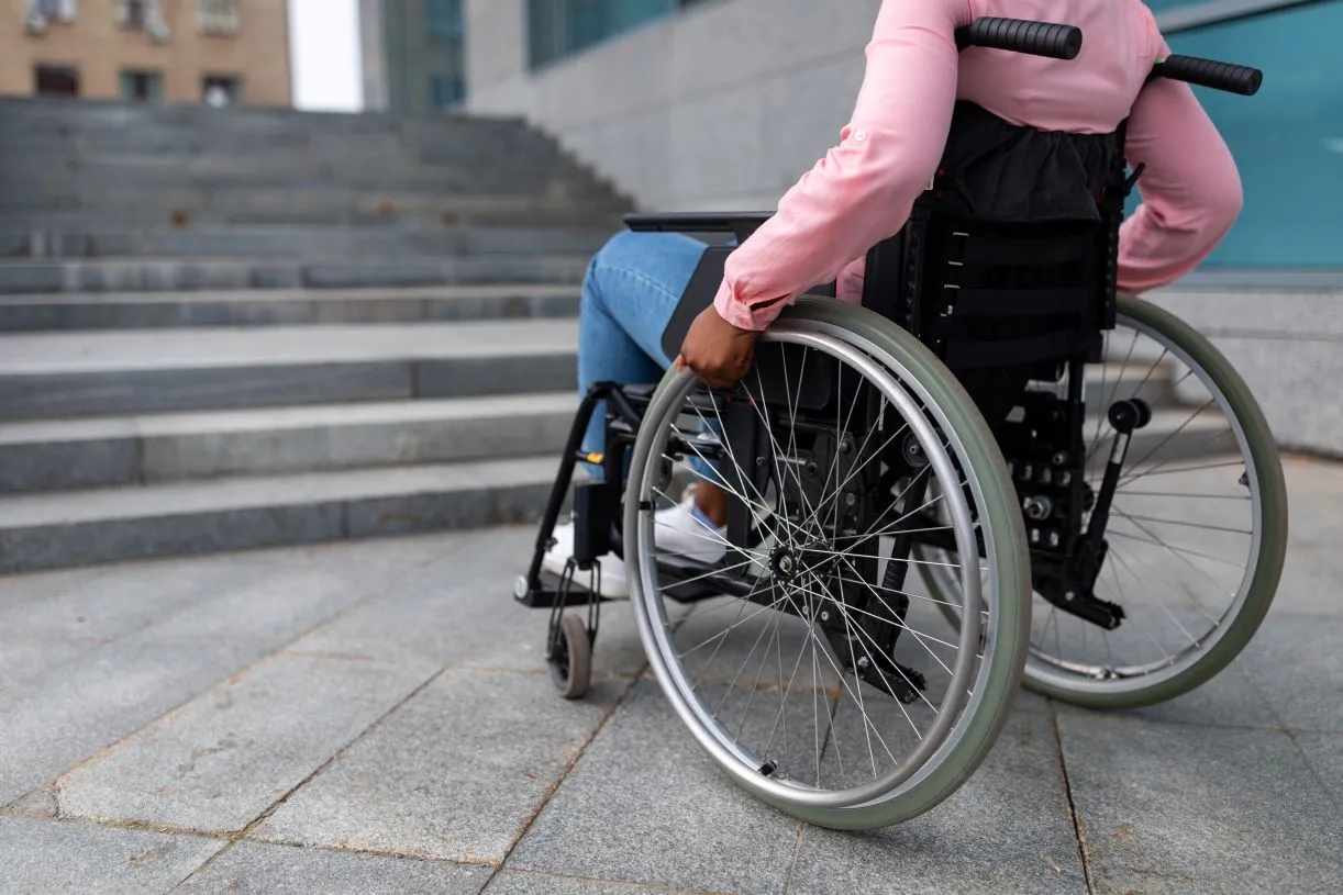 Osoba poruszająca się na wózku inwalidzkim stoi przed schodami, w pobliżu nie widać rozwiązania, które pozwoliłoby się jej przemieścić.