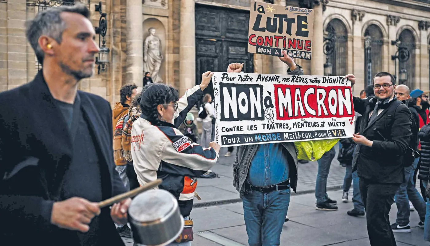Grupa demonstrantów protestuje na ulicy we Francji. Na pierwszym planie mężczyzna uderza w garnek, a w tle inni trzymają transparent z napisem „NON À MACRON” sprzeciwiający się polityce prezydenta.