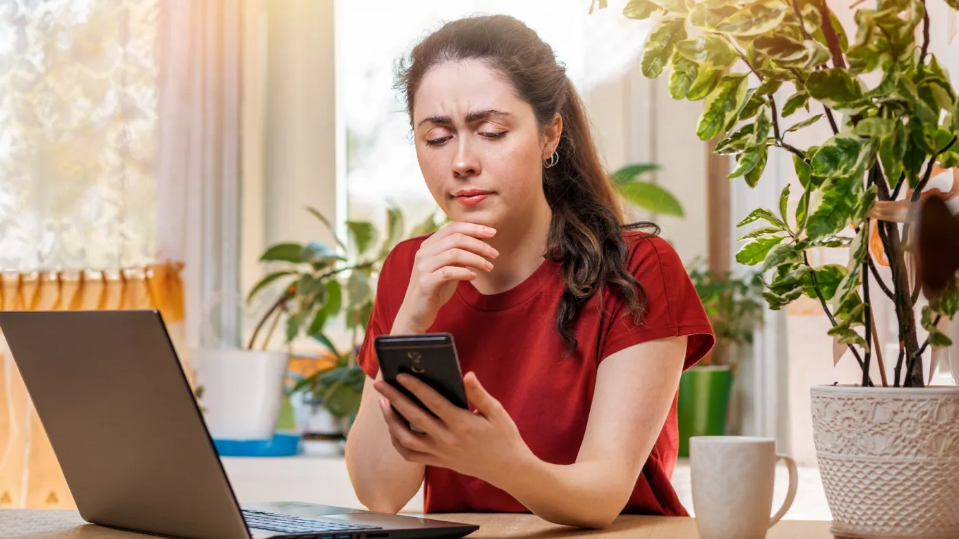 Zmartwiona kobieta siedzi za biurkiem w domu przy laptopie i patrzy w ekran smartfona