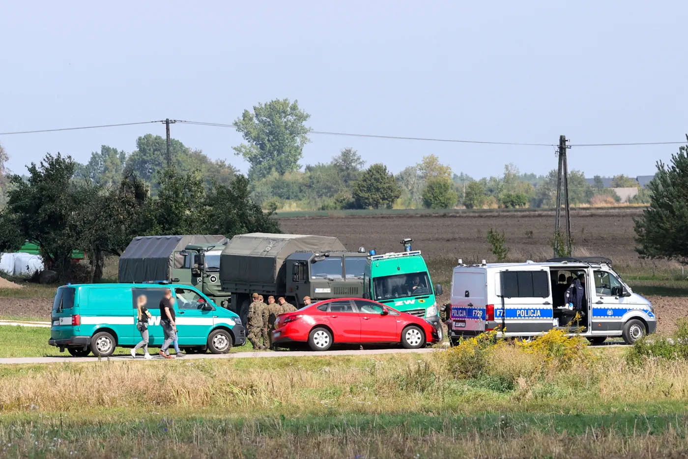 Wojsko i policja przy pojazdach na wiejskiej drodze, w tle pola i drzewa – akcja służb w terenie”