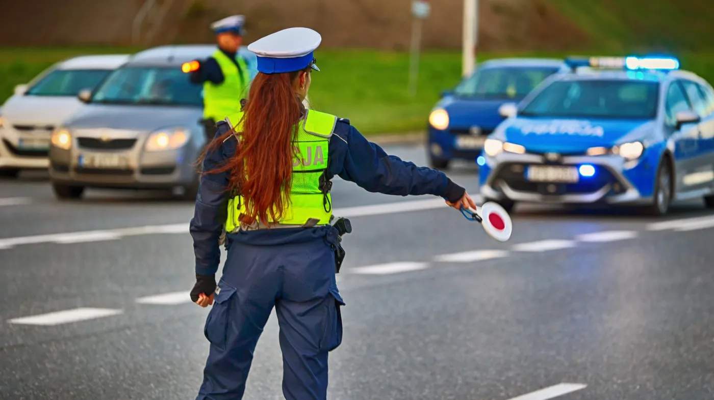 Policjantka drogówki zatrzymuje pojazdy, w tle widać drugiego policjanta, radiowóz i 3 samochody