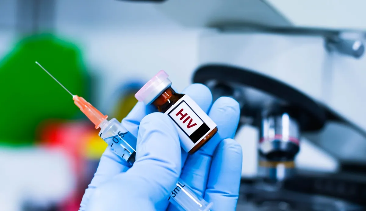 Zbliżenie na dłoń w niebieskiej rękawiczce medycznej, trzymającą szklaną ampułkę z wyraźnym, czarnym napisem "HIV" oraz przezroczystą strzykawkę z igłą. Tło jest jasne, rozmyte, sugerujące wnętrze gabinetu lekarskiego