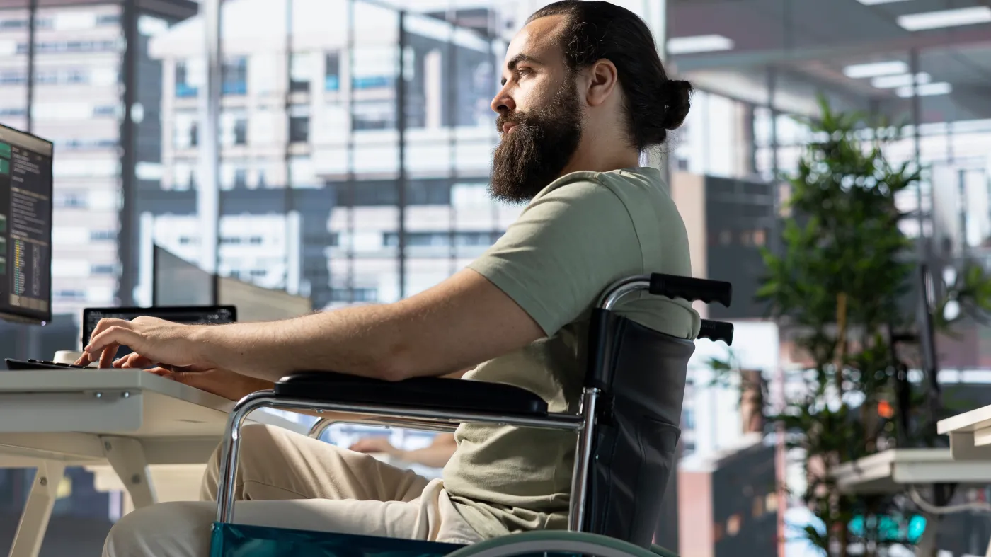 Pracownik z niepełnosprawnością siedzący na wózku w biurze pracujący przy biurkach komputerowych